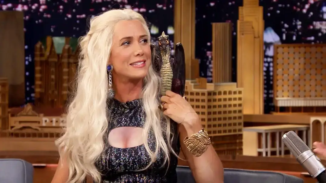Kristen Wiig wears a long blonde wig and a shimmering dress while holding a toy dragon on her shoulder during a late-night talk show appearance.