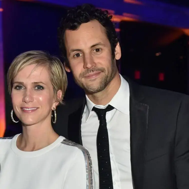 Kristen Wiig, in a white long-sleeved dress with silver detailing, stands next to her husband, Avi Rothman, who is wearing a black suit and tie.