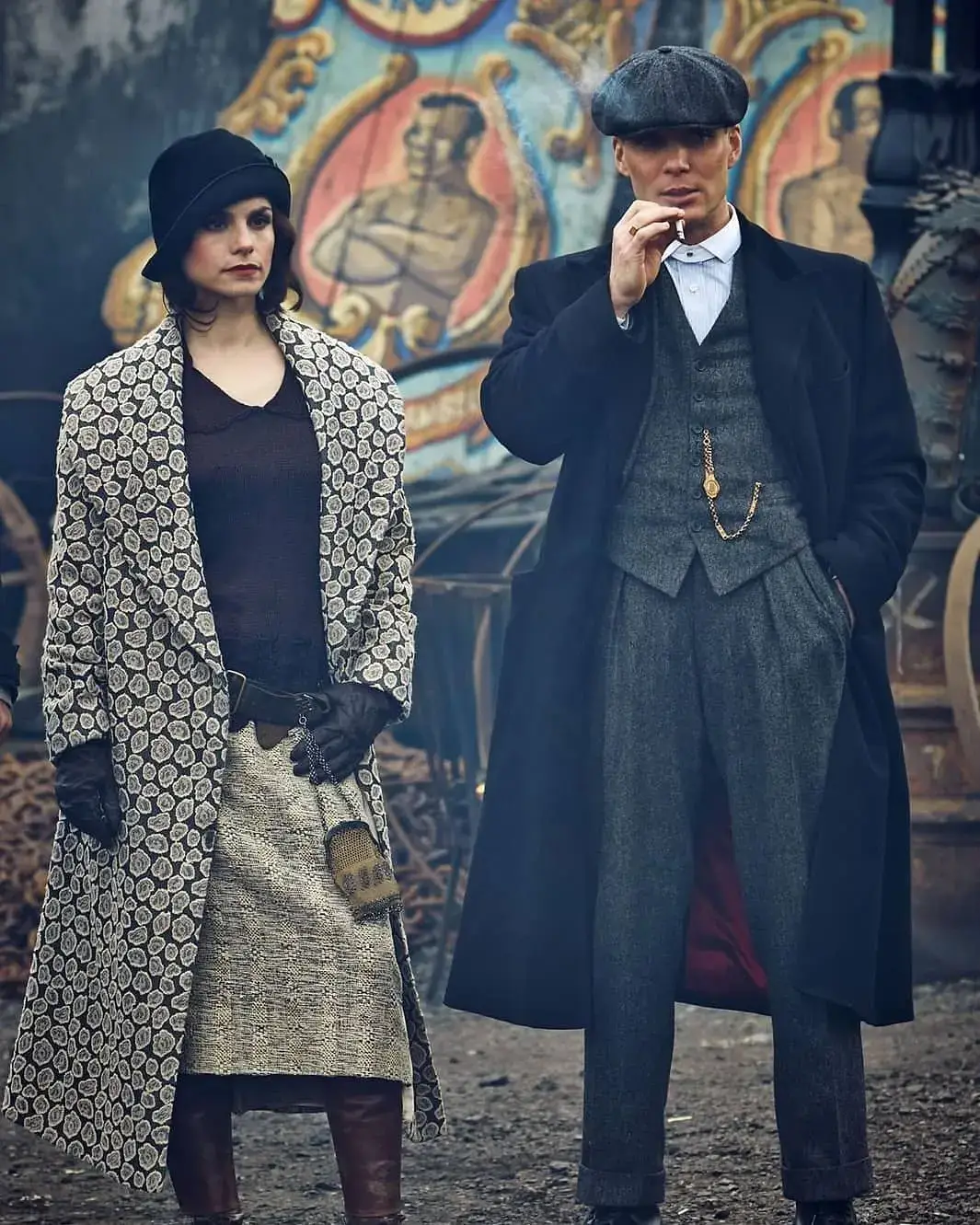 Charlotte Riley as May Carleton in a patterned long coat and hat stands beside Cillian Murphy as Tommy Shelby, who wears a dark overcoat and flat cap while smoking a cigarette.