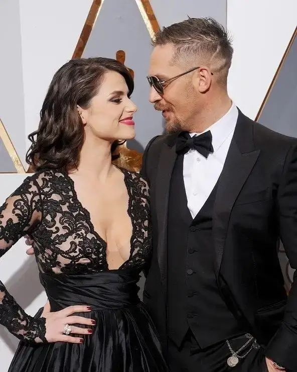 Charlotte Riley, in a black lace gown, and her husband Tom Hardy, in a black tuxedo and sunglasses, look at each other and smile while posing at an awards event.