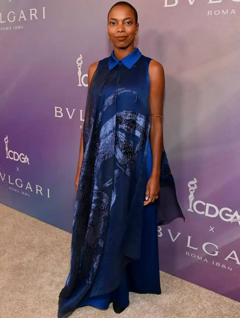 Sasheer Zamata poses on a purple red carpet wearing a sleeveless, flowing blue patterned gown with a collared neckline.