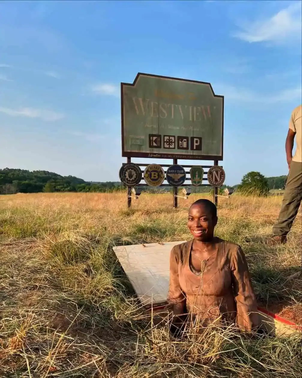 Sasheer Zamata smiles while emerging from a trapdoor in a grassy field in front of a welcome sign for Westview.