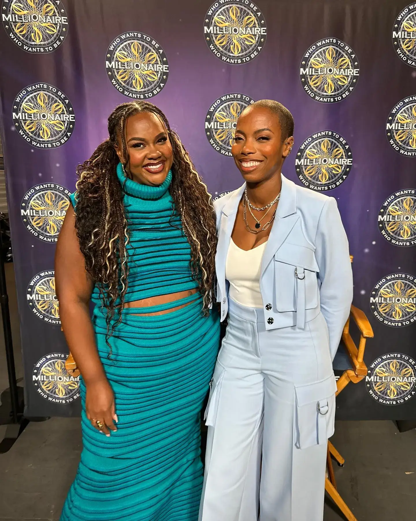 Nicole Byer and Sasheer Zamata pose together in front of a "Who Wants to Be a Millionaire" backdrop wearing a teal ribbed dress and a light blue utility suit, respectively.