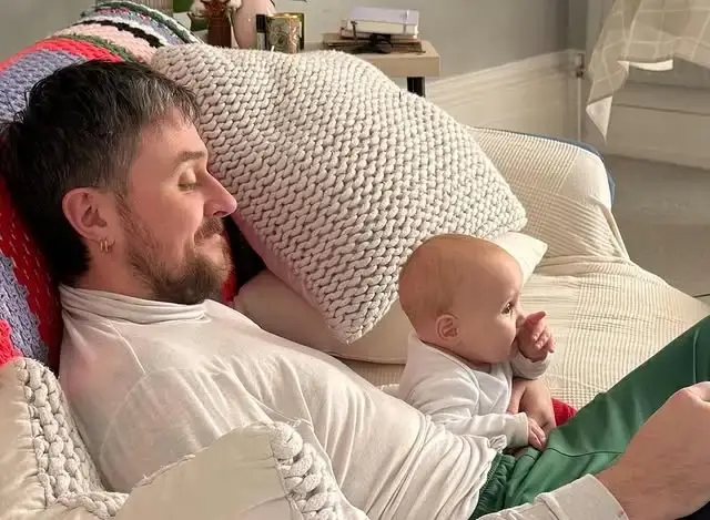 Sam C Wilson relaxes on a sofa with a colorful knit blanket while a young baby sits beside him.