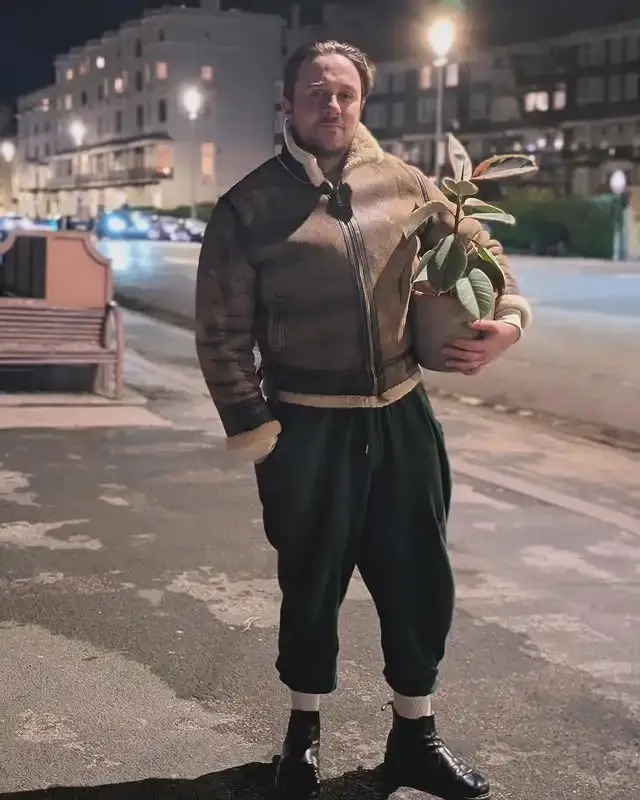Sam C Wilson stands on a city sidewalk at night wearing a shearling jacket and dark trousers while holding a potted plant.