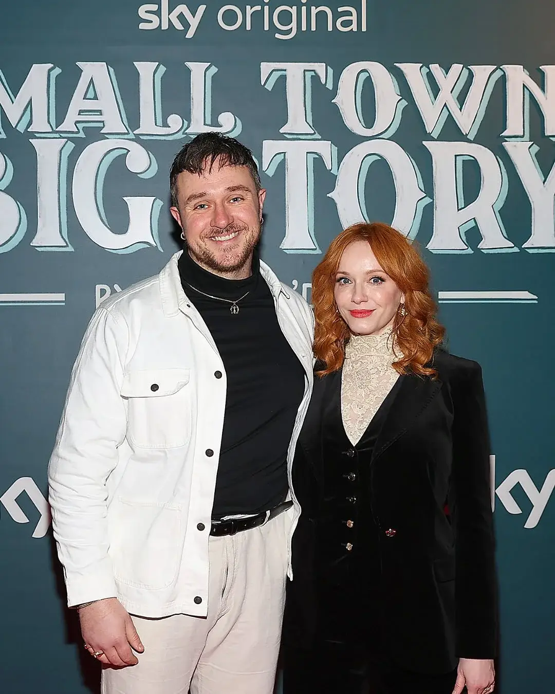 Sam C Wilson and Christina Hendricks smile together on a red carpet in front of a teal "Small Town, Big Story" backdrop.