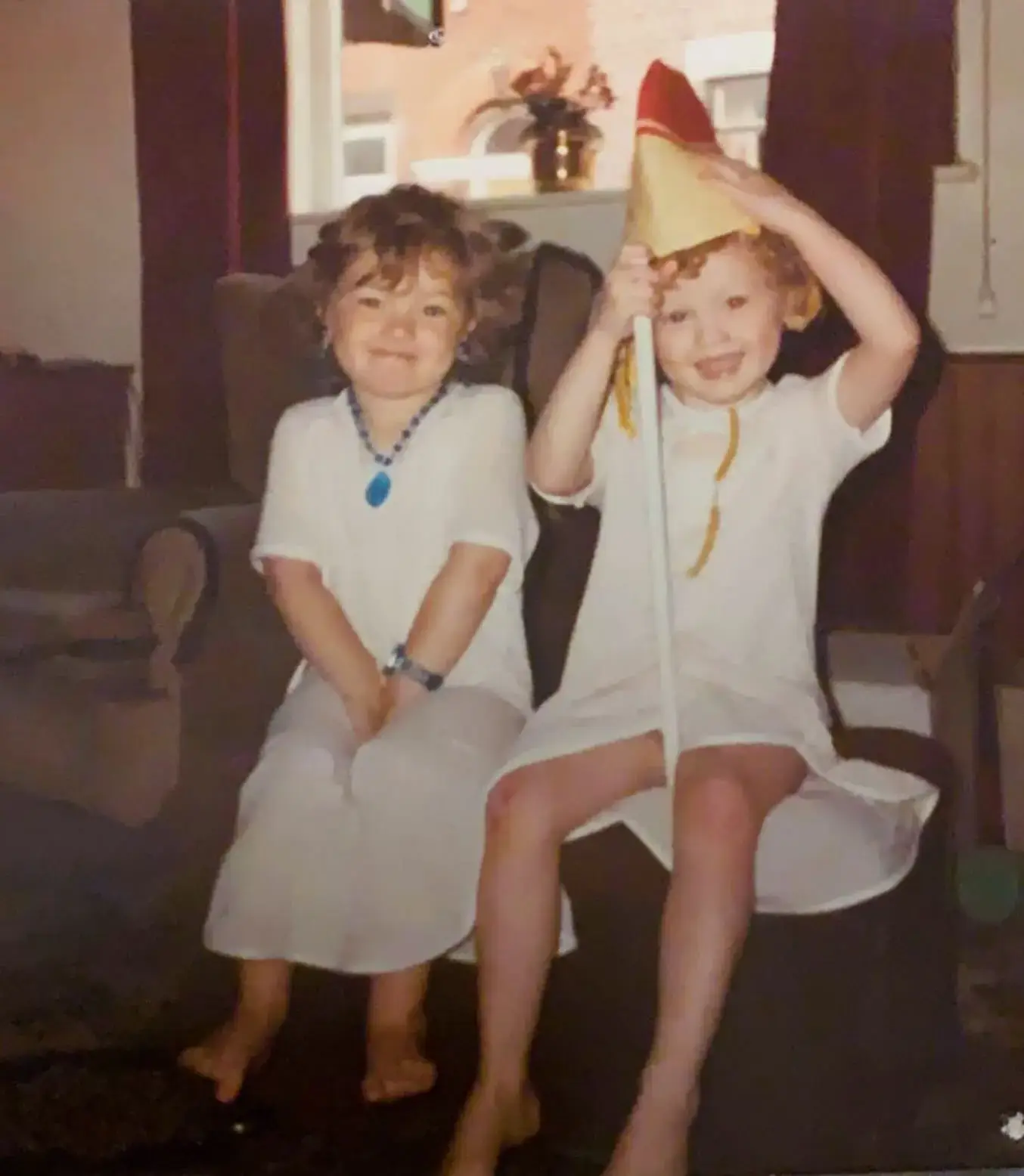 A vintage childhood photo shows Karen Elson on the right holding a toy flagpole and her sister on the left wearing a blue necklace as they sit together indoors.