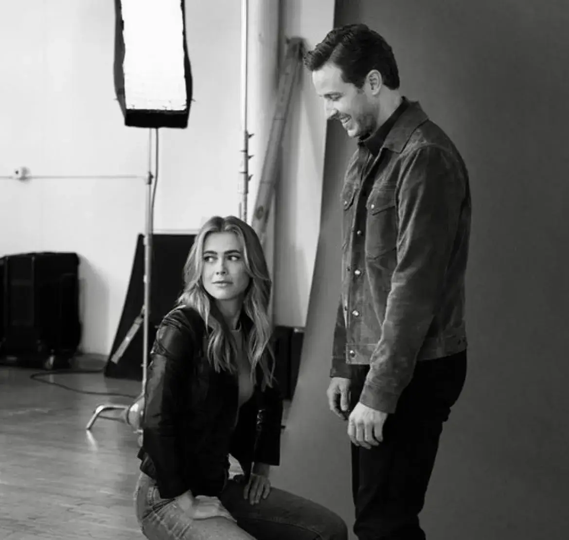 A black and white photo of Melissa Roxburgh sitting on the floor looking back at a smiling Josh Dallas who is standing next to her in a studio setting.