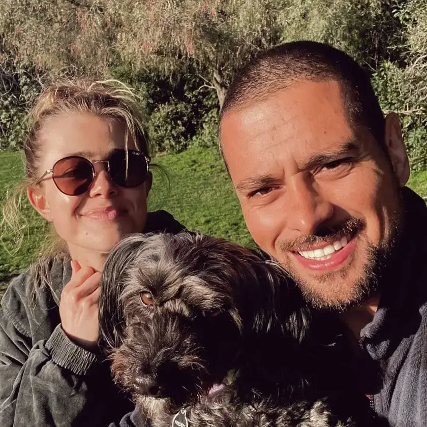 Melissa Roxburgh and J.R. Ramirez smile for an outdoor selfie while posing with a small, shaggy black dog in a sunny park setting.
