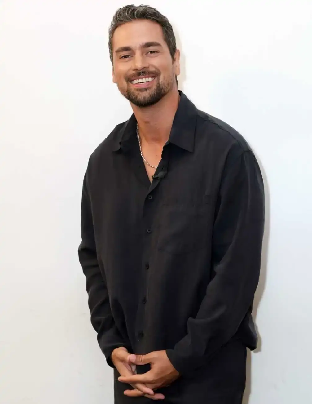 J.R. Ramirez stands against a plain white wall, smiling broadly while wearing a black button-down shirt and a silver chain.
