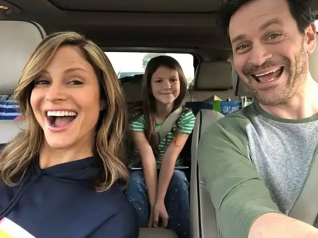 Andrea Savage, Jeremy Plager, and their daughter smile broadly while taking a bright family selfie inside a car.