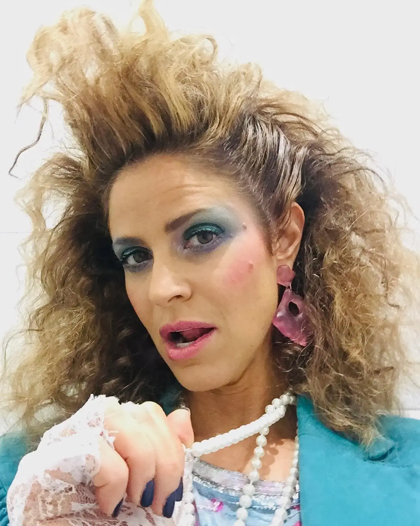 ndrea Savage poses with voluminous 1980s-style hair, bold blue eyeshadow, and a lace glove while wearing a turquoise blazer and pearls.