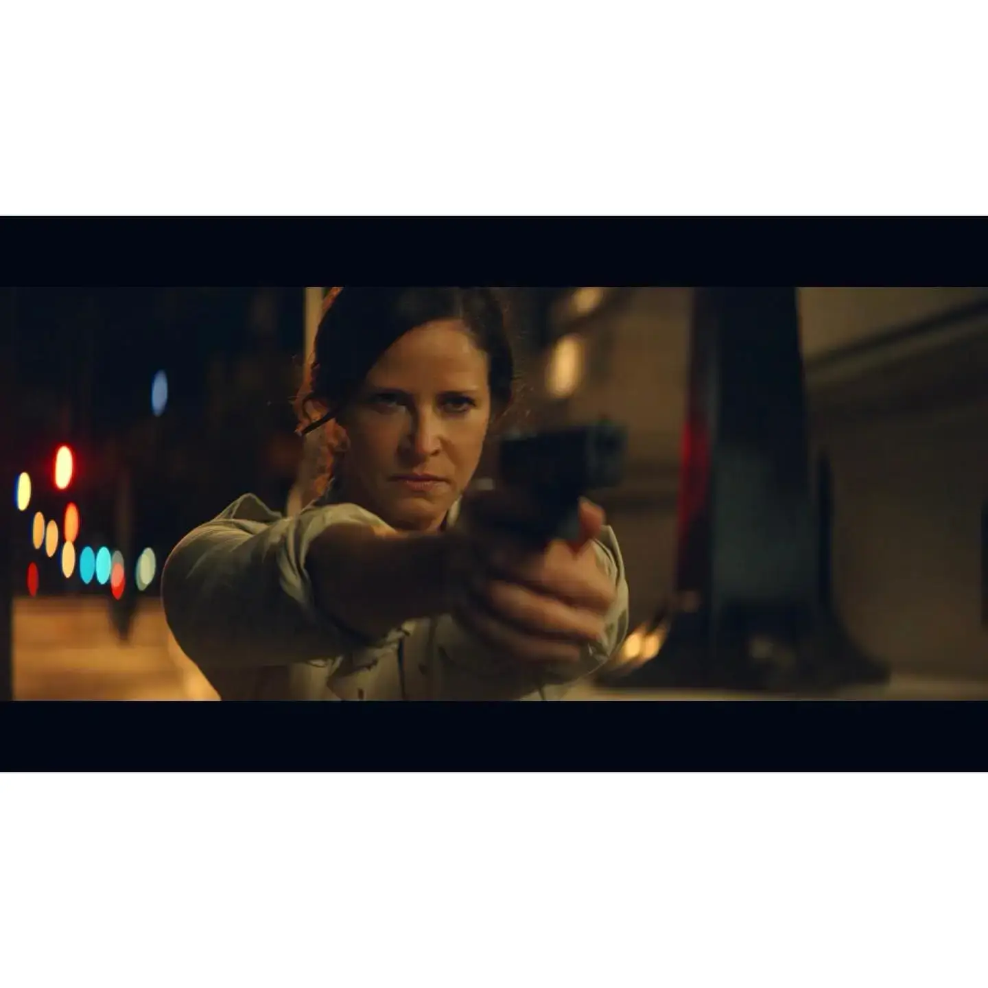 Andrea Savage aims a handgun directly at the camera with a determined expression in a dimly lit, cinematic night scene.