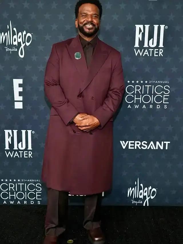 Actor Craig Robinson poses on a red carpet wearing a long, double-breasted maroon coat over a dark shirt and trousers.