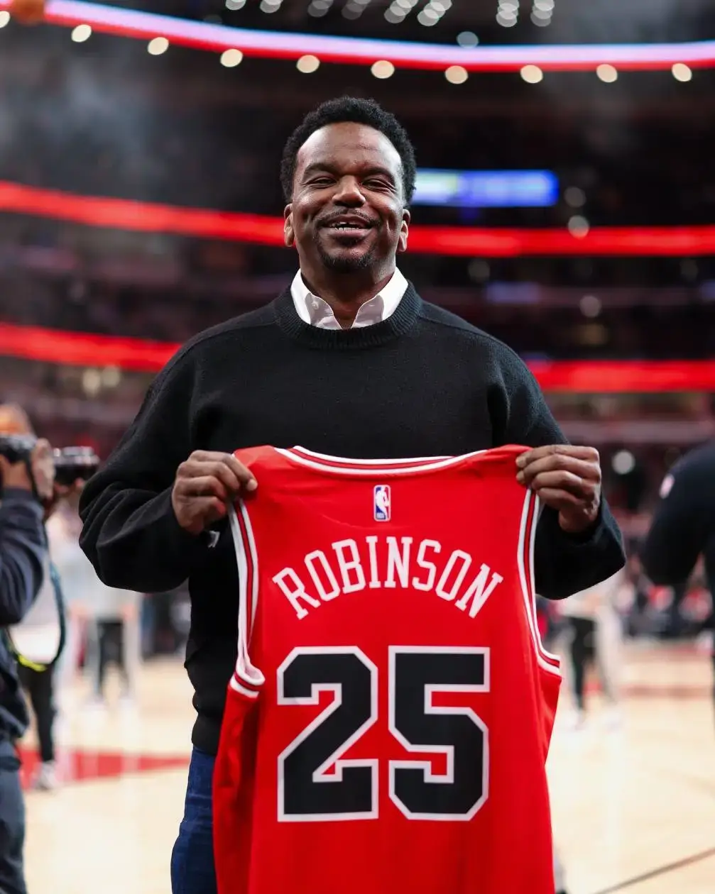 Actor Craig Robinson smiles while holding up a red Chicago Bulls jersey featuring his name and the number 25 at a basketball arena.