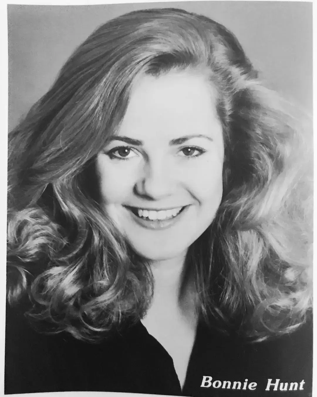 A black and white portrait of Bonnie Hunt smiling with her long, wavy hair flowing around her shoulders.