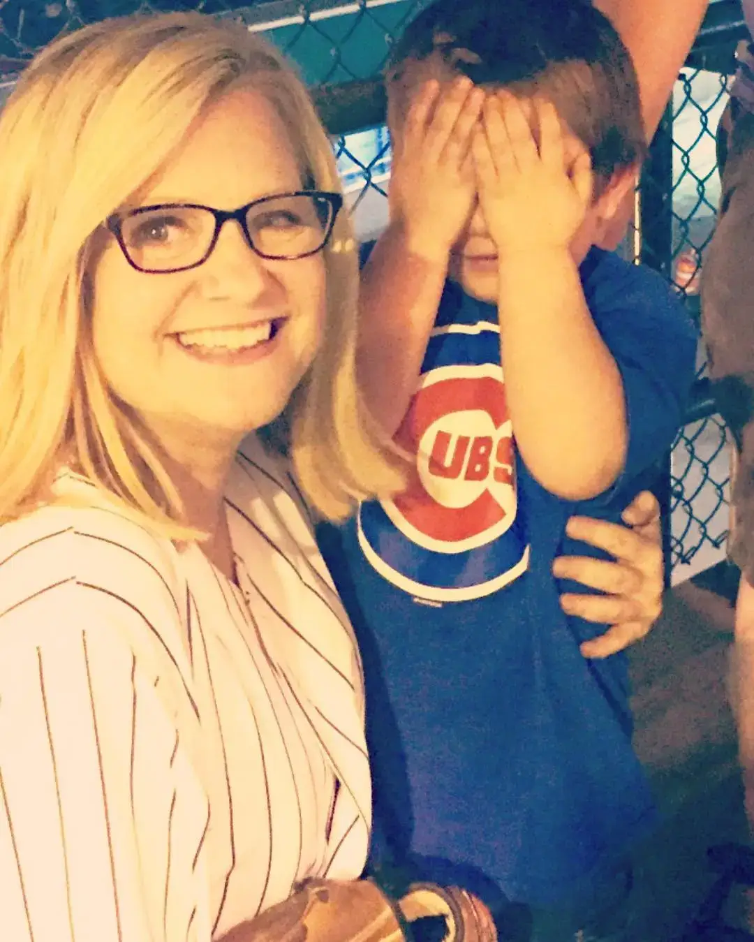Bonnie Hunt smiles broadly while standing next to a young child in a blue Chicago Cubs shirt who is playfully covering their eyes with their hands.