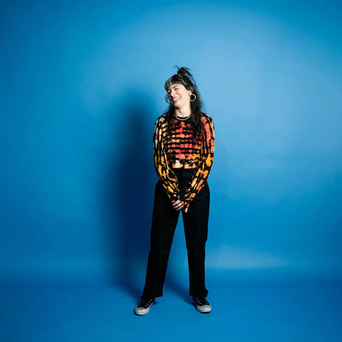 Melissa Villaseñor stands centered against a solid blue backdrop, laughing while wearing a vibrant orange and black tie-dye shirt and black trousers.