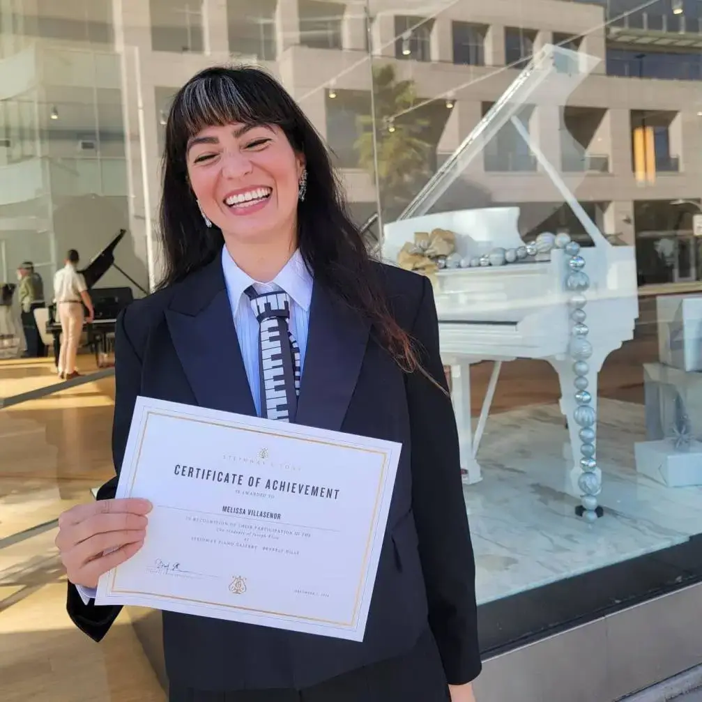 Melissa Villaseñor wears a suit and piano-key tie while holding a "Certificate of Achievement" from Steinway & Sons.
