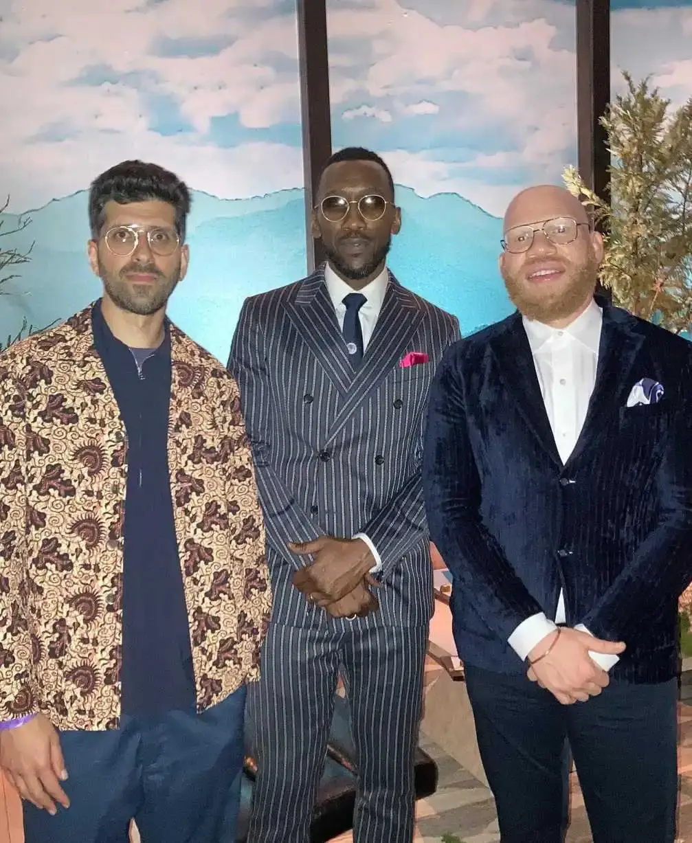 Barmak Badaei, Mahershala Ali, and Krondon stand side-by-side in stylish suits and patterns against a blue mountain backdrop.