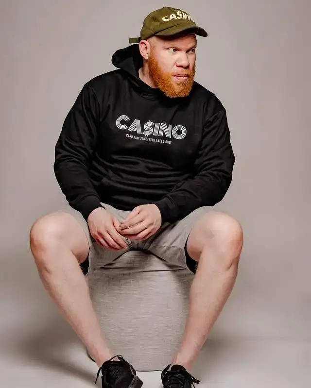 Krondon sits on a stool wearing a black "CASINO" hoodie, an olive green cap, and grey shorts.