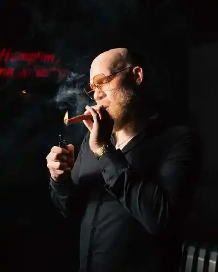 Krondon, wearing a black shirt and yellow-tinted glasses, lights a cigar in a dark room.