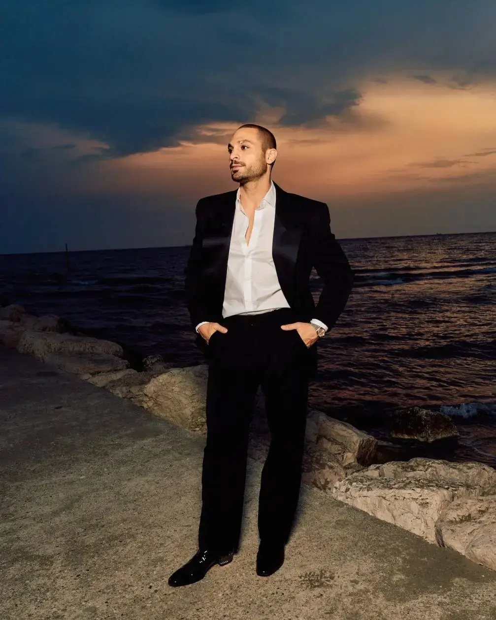 Michael Mando poses in a black suit on a rocky coastline during a sunset.