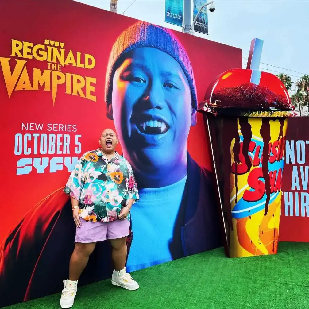 acob Batalon poses in a floral shirt and purple shorts in front of a large "Reginald the Vampire" promotional billboard.