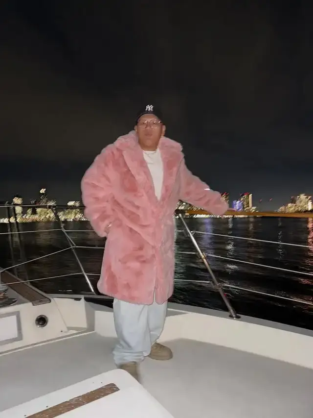 Jacob Batalon wears a large pink faux-fur coat while standing on a boat deck at night.