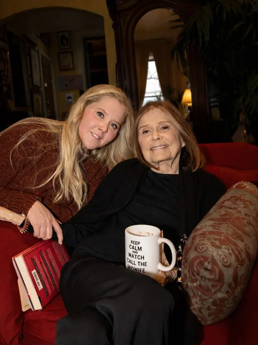 Amy Schumer and Gloria Steinem sit together on a red sofa while holding a book and a "Call the Midwife" mug.