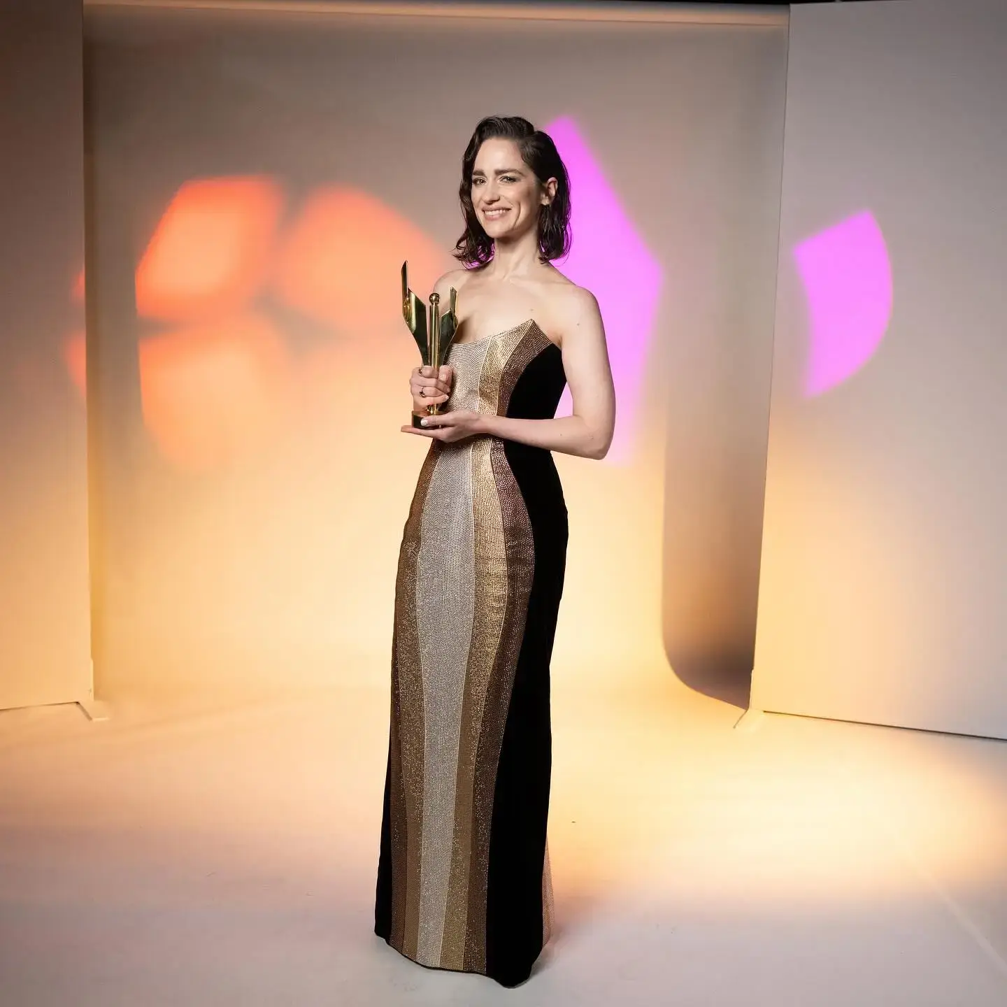 Actress Melanie Scrofano poses in a strapless gold and black gown while holding an award against a brightly lit backdrop.