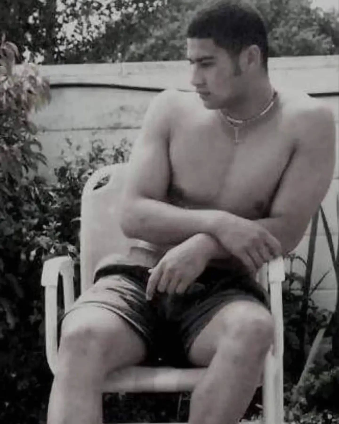 A black and white vintage photo of a young John Tui sitting shirtless in a white plastic chair outdoors.