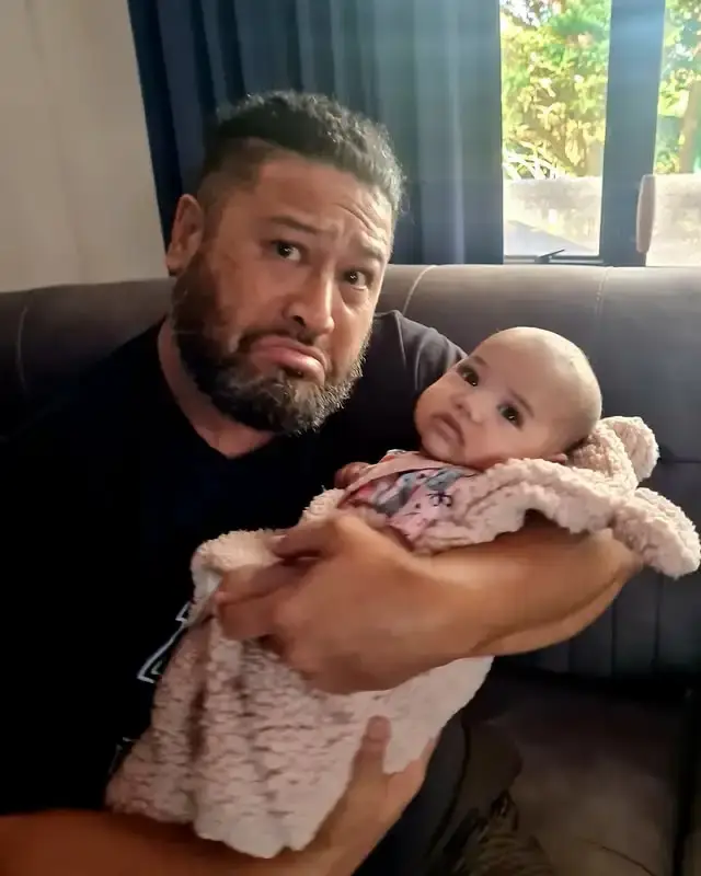 John Tui makes a playful, pouting expression while cradling his baby wrapped in a soft pink blanket.