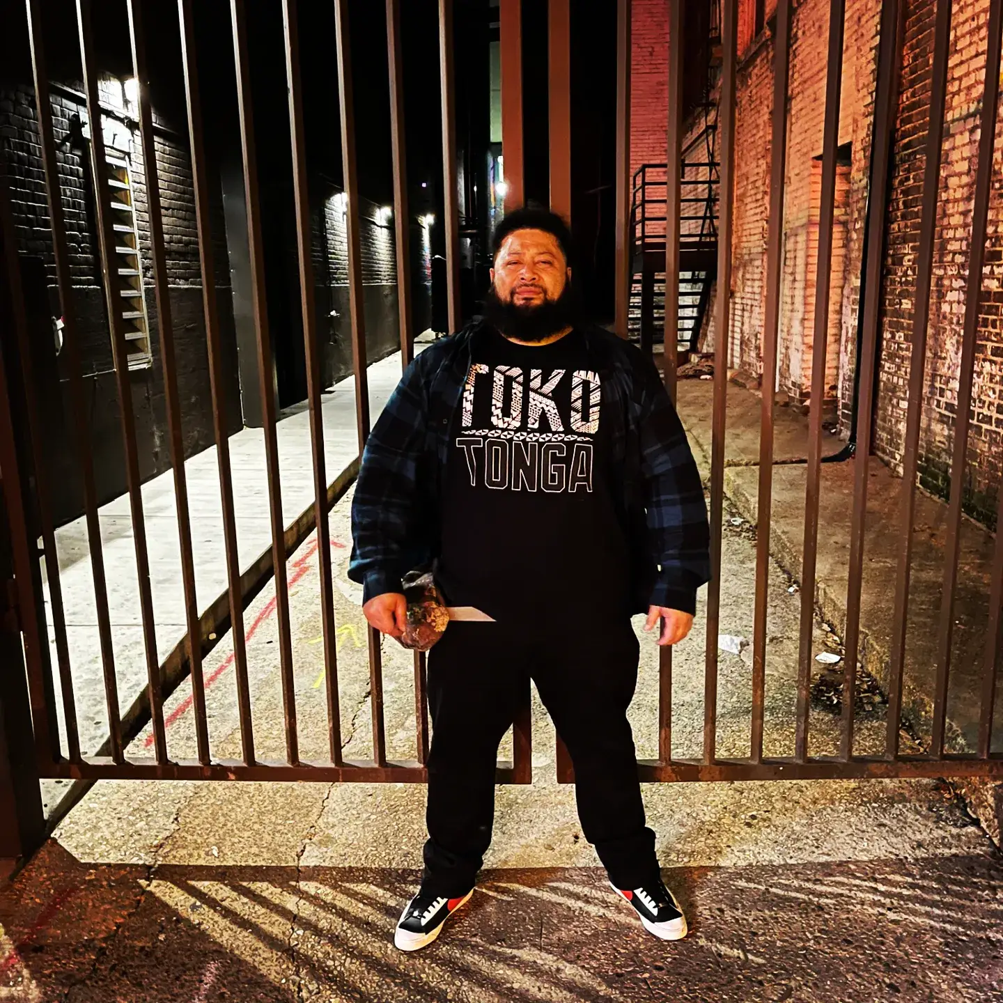 John Tui stands in front of a tall metal gate at night, wearing a "Toko Tonga" t-shirt and a flannel shirt.