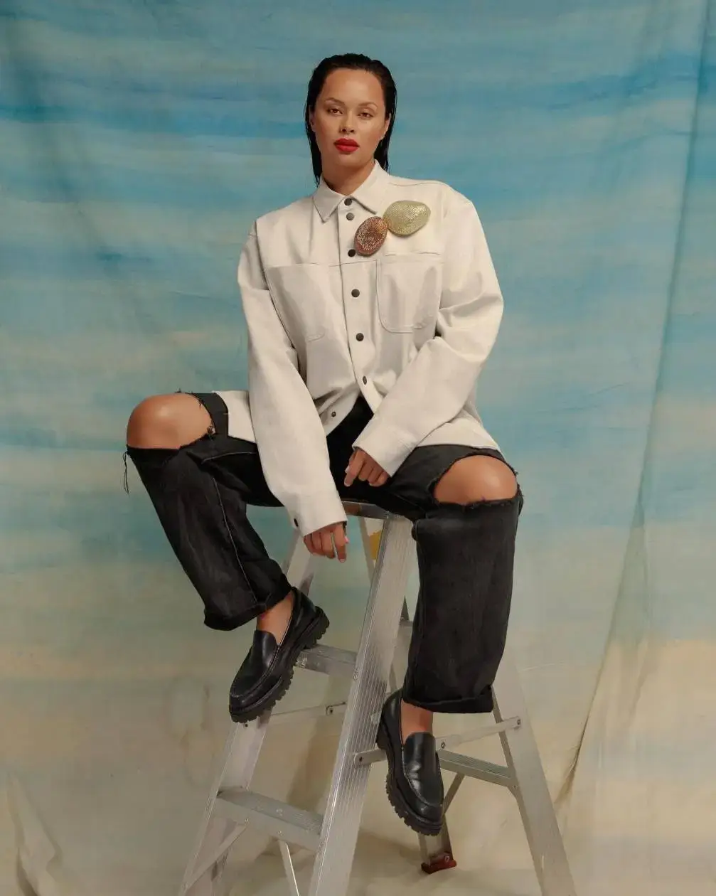 Actress Frankie Adams poses while seated on a silver stepladder against a blue and beige painted backdrop.