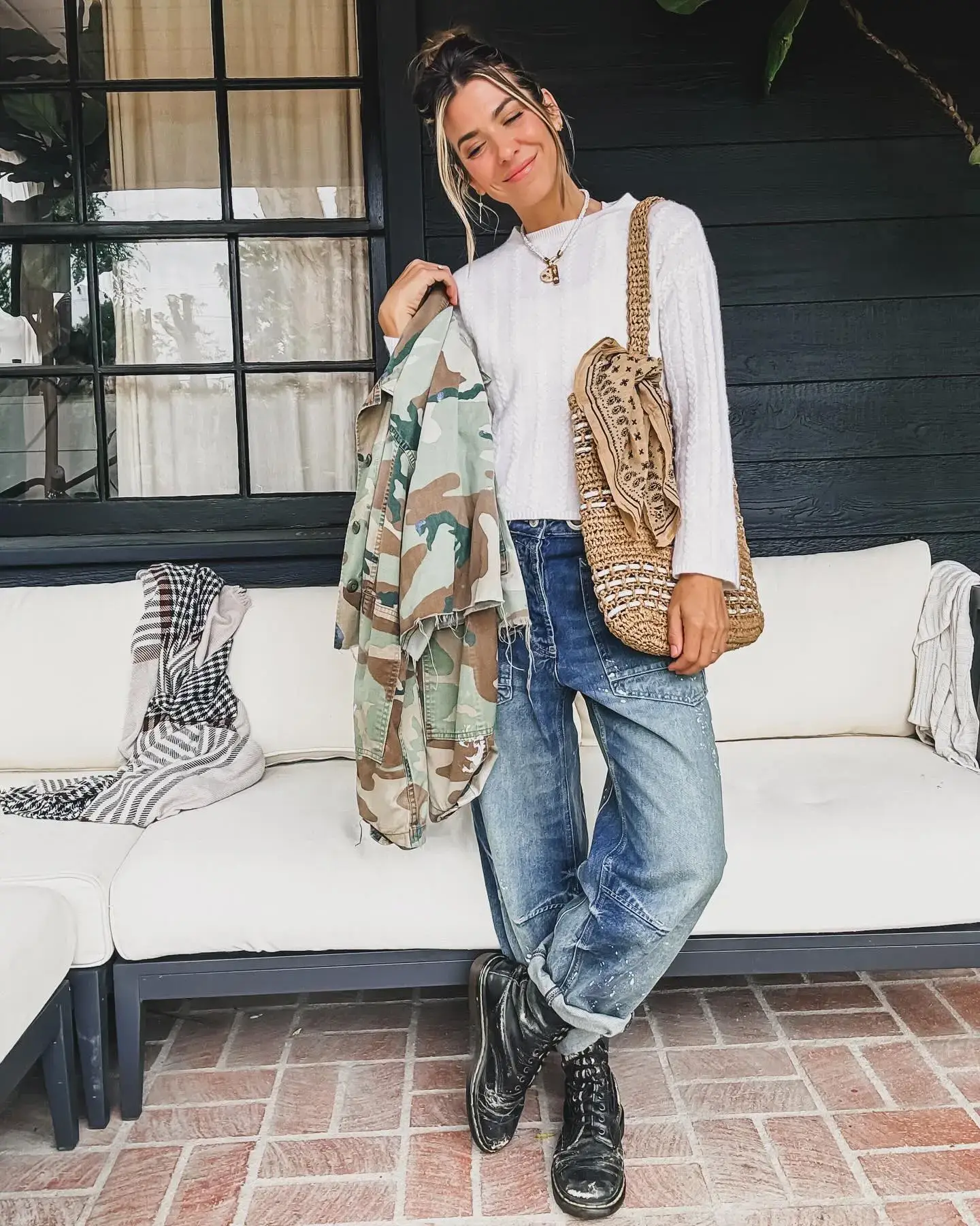 Orly Shani smiles while posing in a white sweater, baggy jeans, and a camouflage jacket on a patio.