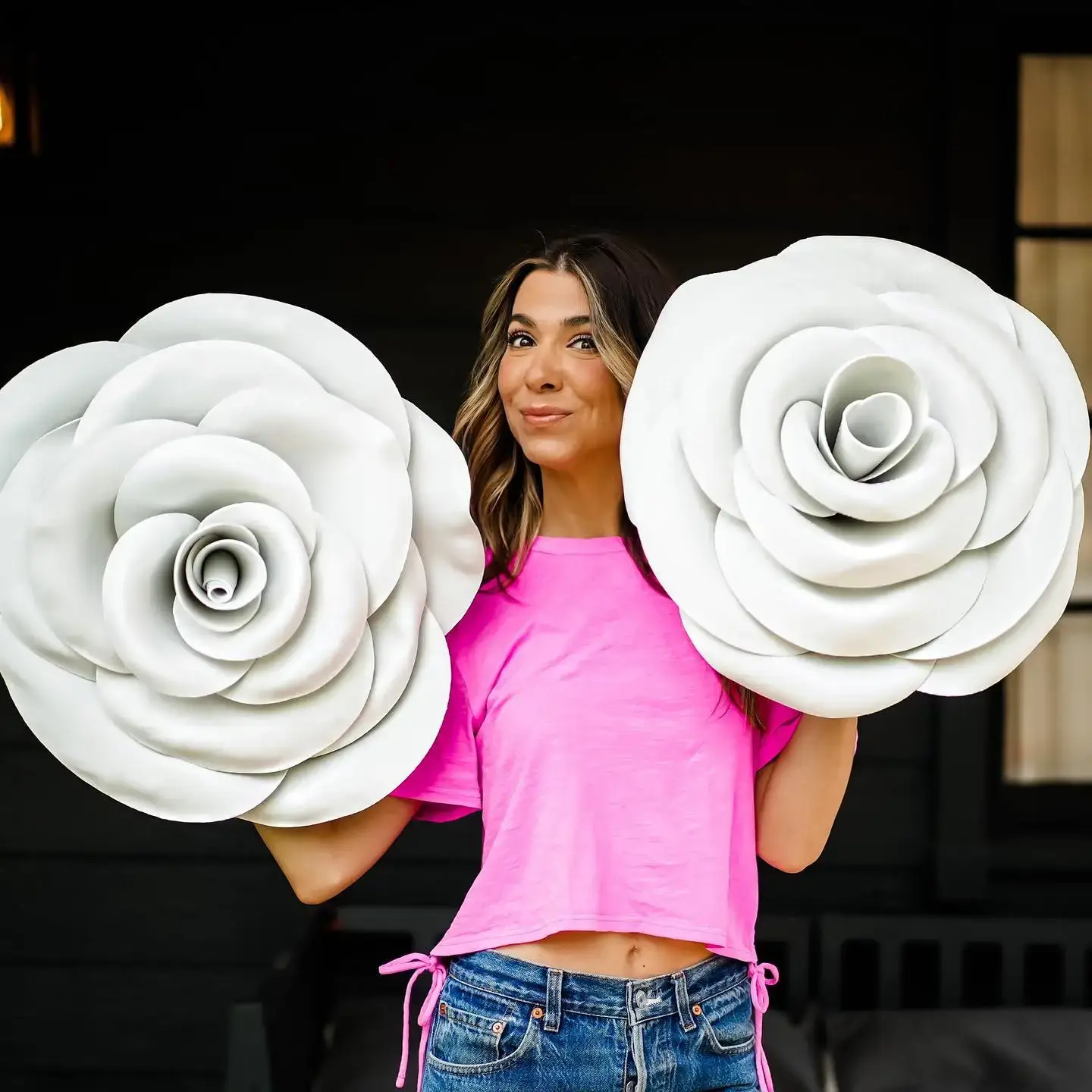 Orly Shani poses in a bright pink t-shirt and jeans while holding two oversized white decorative roses.