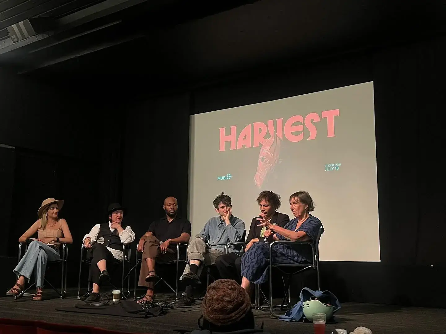 Actor Frank Dillane sits on a panel stage next to colleagues in front of a large screen projecting the title "HARVEST."