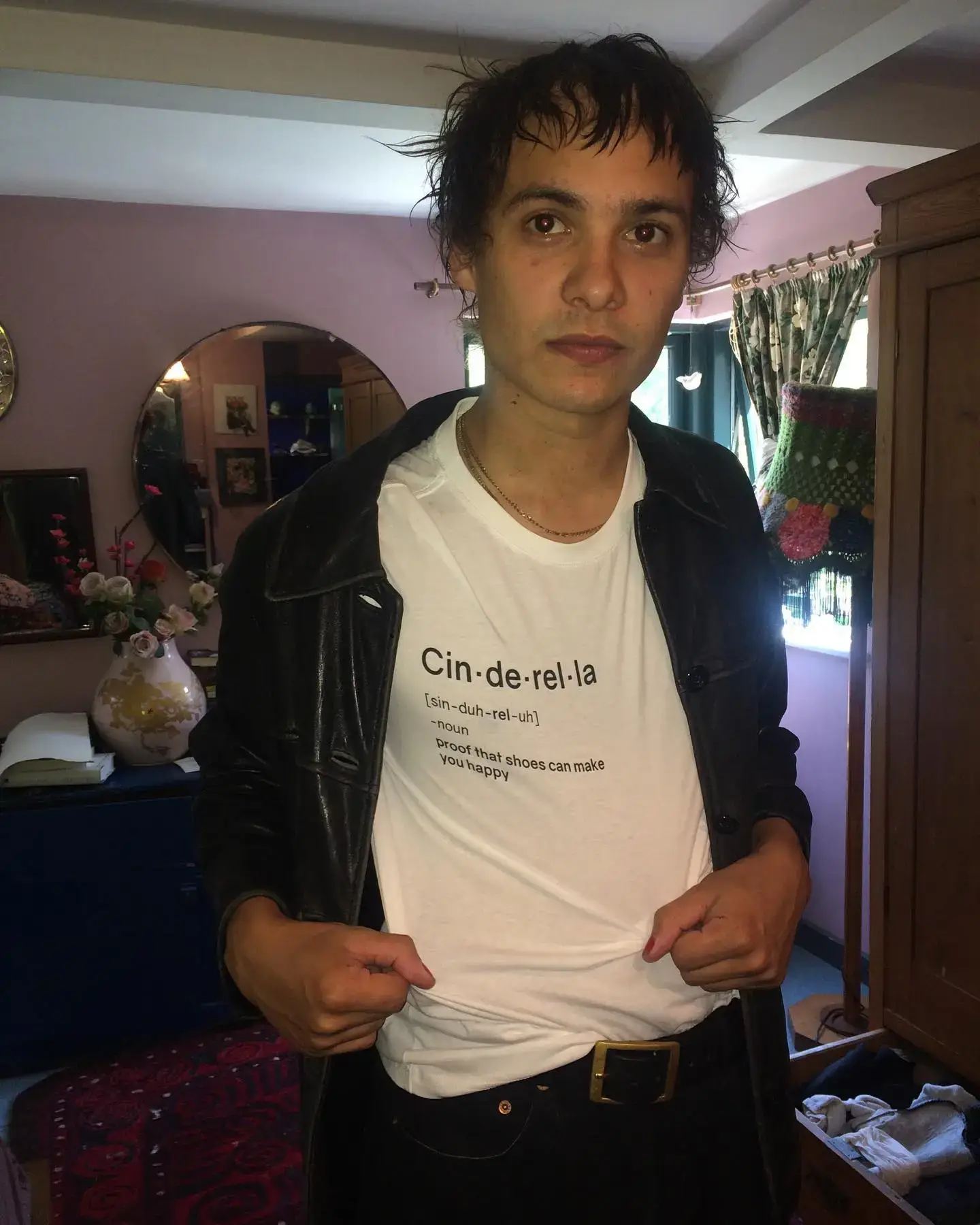 Frank Dillane stands in a room wearing a leather jacket and a white t-shirt featuring a dictionary-style definition of "Cinderella."