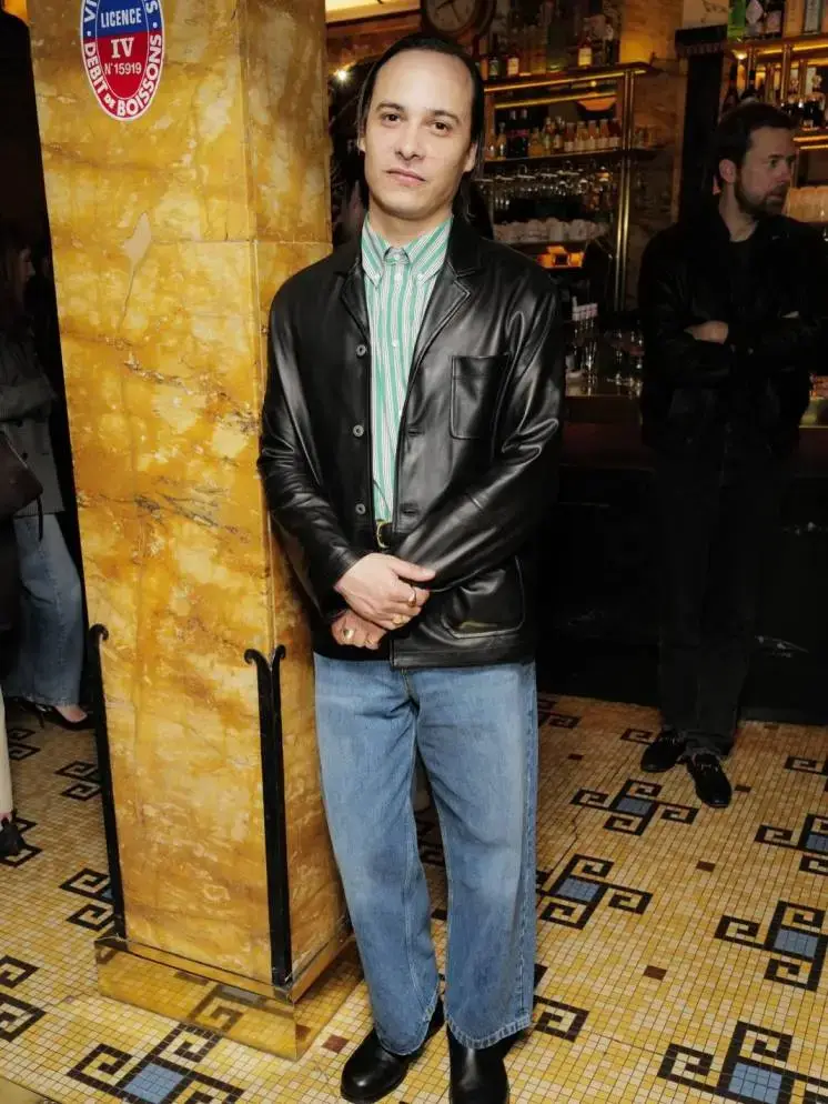 Actor Frank Dillane stands with hands clasped in a leather jacket, striped shirt, and blue jeans at an indoor event.