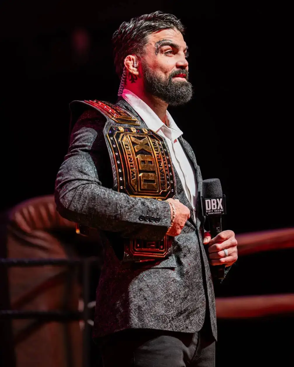 Combat sports star Mike Perry stands proudly in a textured grey blazer, holding the gold and black "DBX" championship belt over his shoulder.