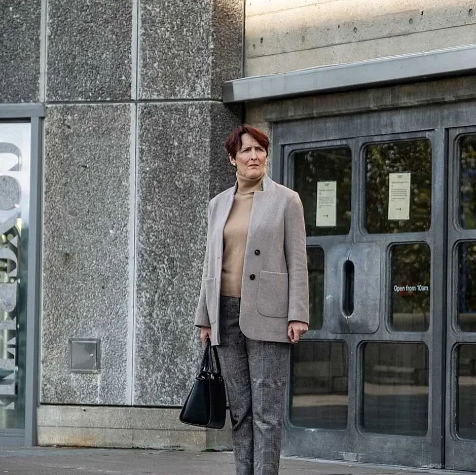Actress Fiona Shaw stands outside a concrete building wearing a tan blazer and patterned trousers while looking off-camera.