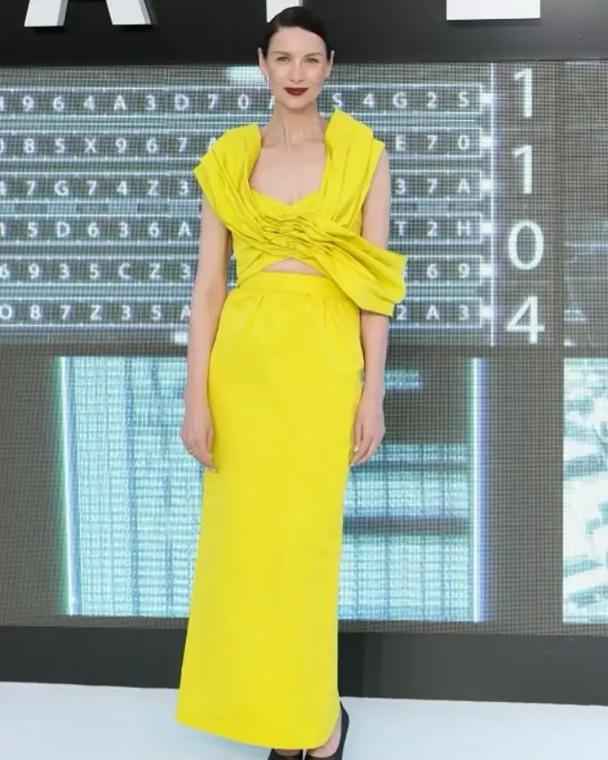 Caitriona Balfe poses on a red carpet wearing a striking, sleeveless bright yellow gown with a draped bodice.
