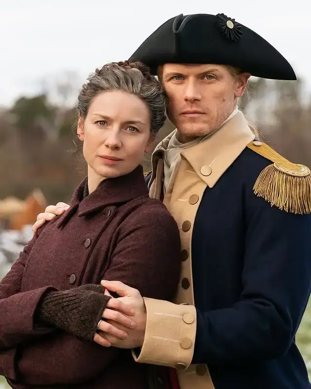 Caitriona Balfe and Sam Heughan stand closely together in 18th-century period costumes against a blurred outdoor background.