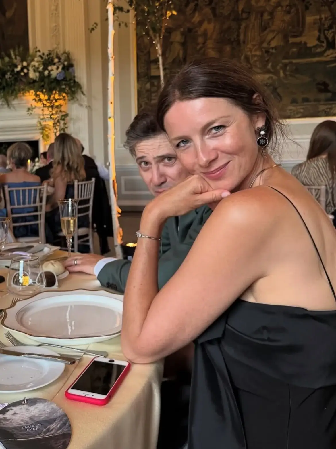 Caitriona Balfe smiles at a dinner table while her husband, Tony McGill, leans in behind her at a formal event.