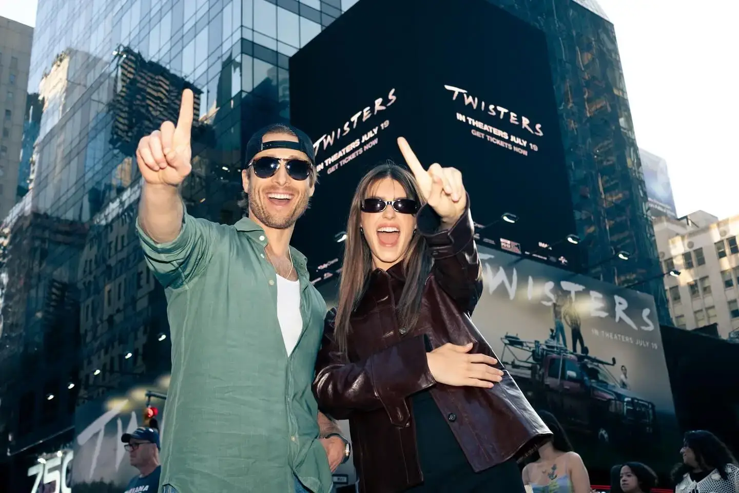 Daisy Edgar-Jones and Glen Powell pose excitedly in front of a large "Twisters" movie billboard in a city.