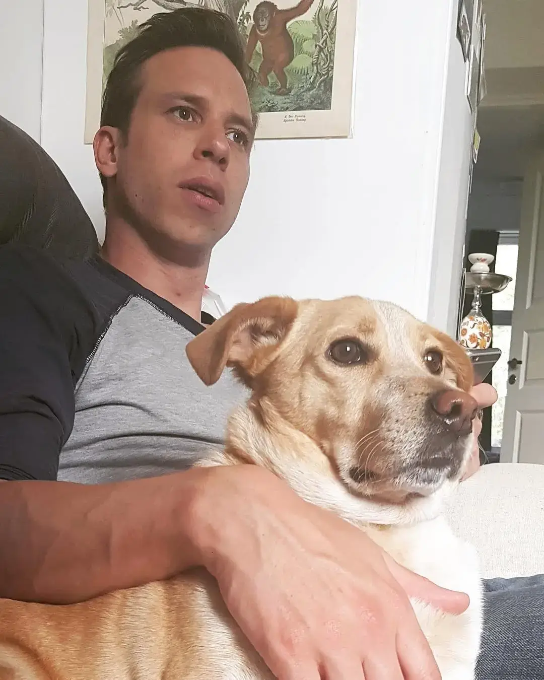 Herbert Nordrum sits on a couch while holding his tan and white dog, Mumi, who is looking off-camera.