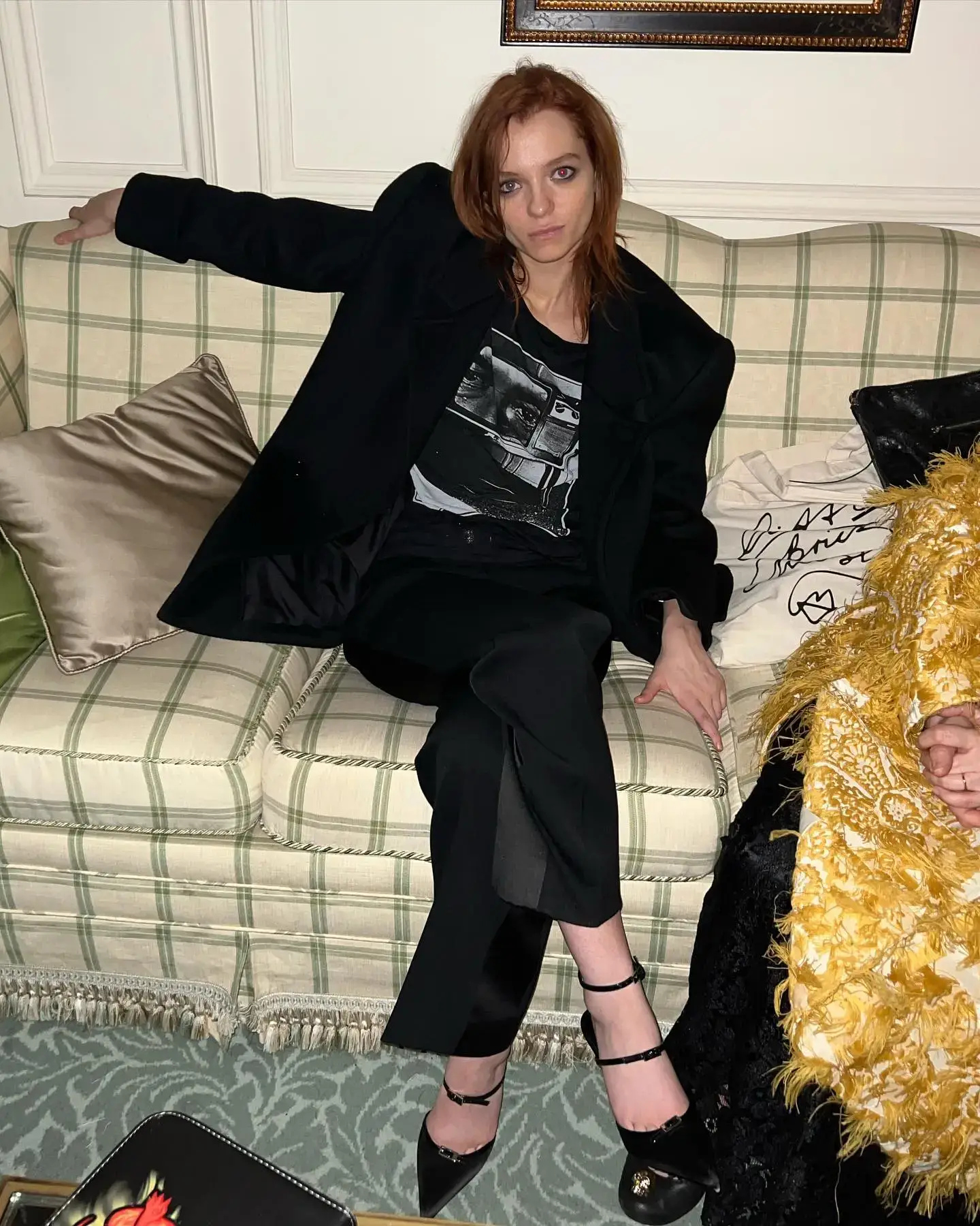 Esmé Creed-Miles sits on a checkered sofa wearing an oversized black coat, a graphic t-shirt, and black pointed heels.