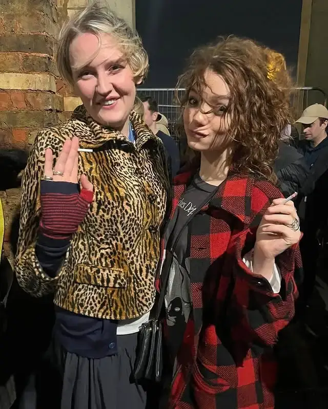 Esmé Creed-Miles stands outdoors wearing a red plaid coat and holding a cigarette while posing next to a woman in a leopard-print jacket.