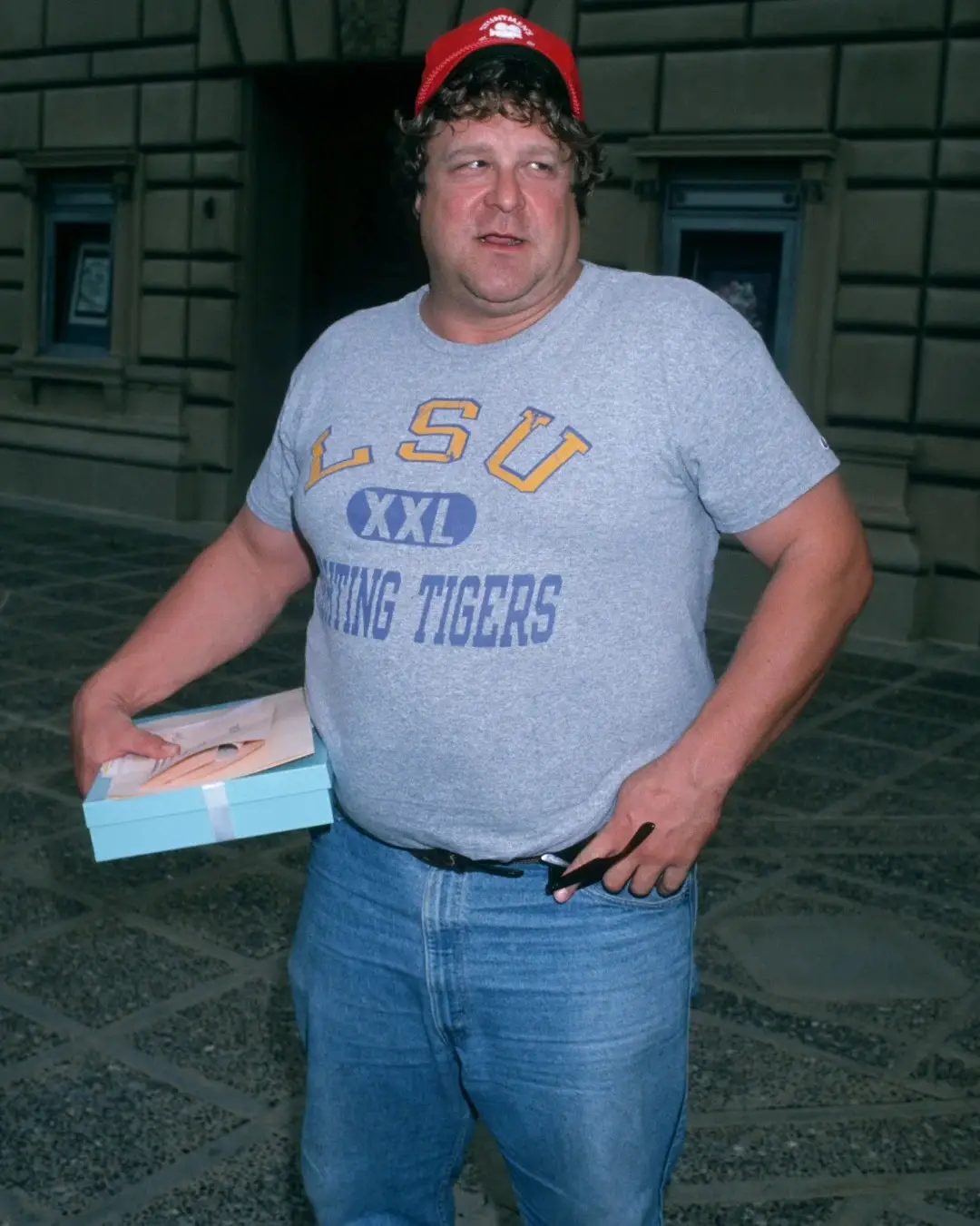 A younger John Goodman stands outdoors wearing a grey LSU t-shirt, blue jeans, and a red baseball cap while holding a small blue box.